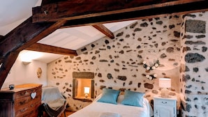Un dormitorio con pared de piedra, vigas de madera, cama, cómoda, silla y mesita de noche con lámpara.
