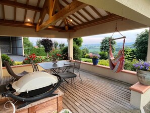 Un patio couvert avec une terrasse en bois, une table et des chaises, un barbecue et un fauteuil suspendu.