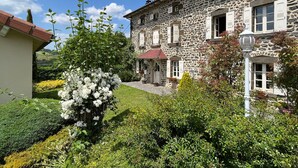 Ein Gebäude aus Stein mit rotem Ziegeldach, ein kleiner Garten mit weißen Blüten und ein klarer, blauer Himmel.