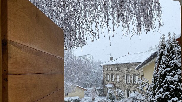 Eine Holztür, die zu einem verschneiten Innenhof mit einem Steinhaus und schneebedeckten Bäumen führt.