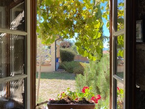 A window with a view of a garden, including a grapevine, a wooden planter with red flowers, and a hedge. - Character house in the heart of the village (Saint-Paulien)