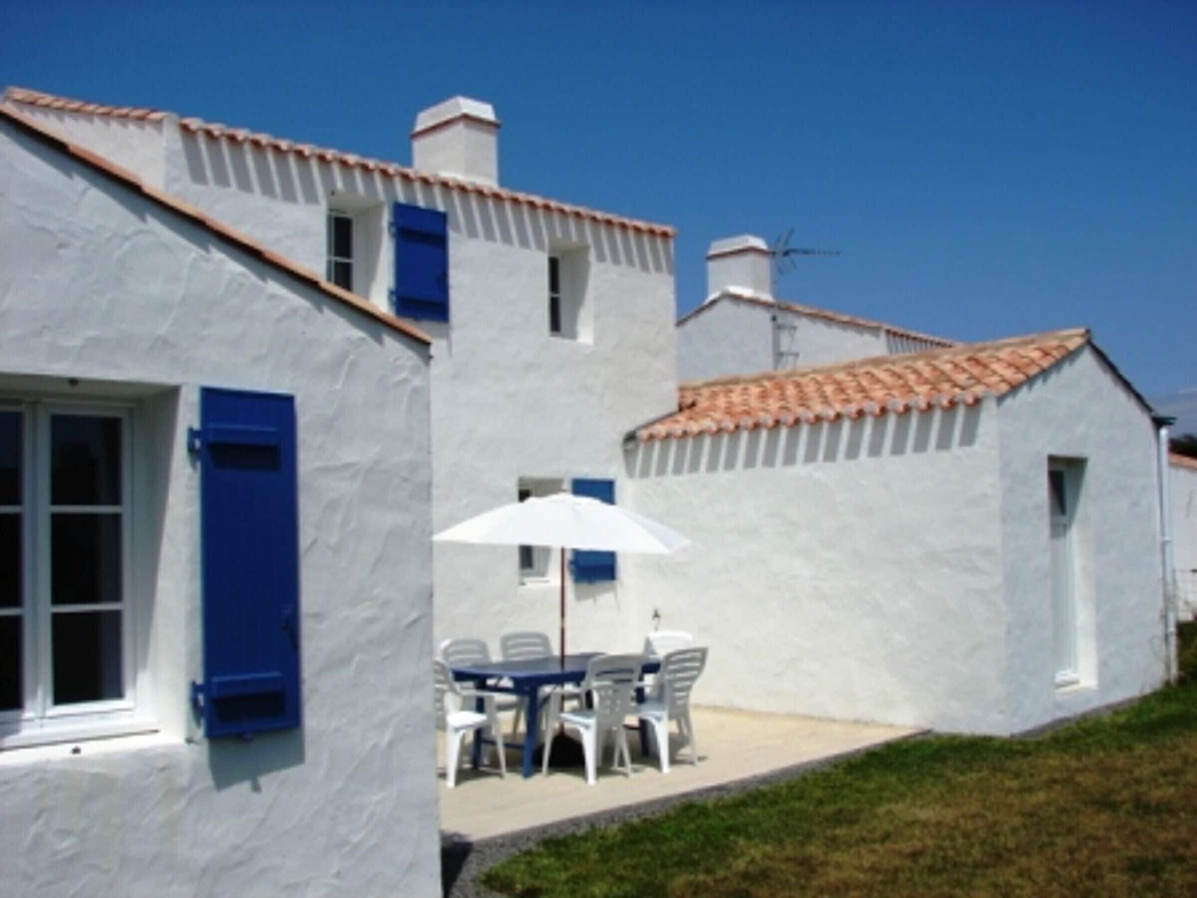 Un bâtiment blanc avec des volets bleus, un toit en tuiles et un petit patio avec une table et des chaises.
