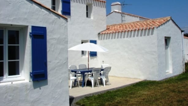 Ein weißes Gebäude mit blauen Fensterläden, einem Ziegeldach und einer kleinen Terrasse mit Tisch und Stühlen.
