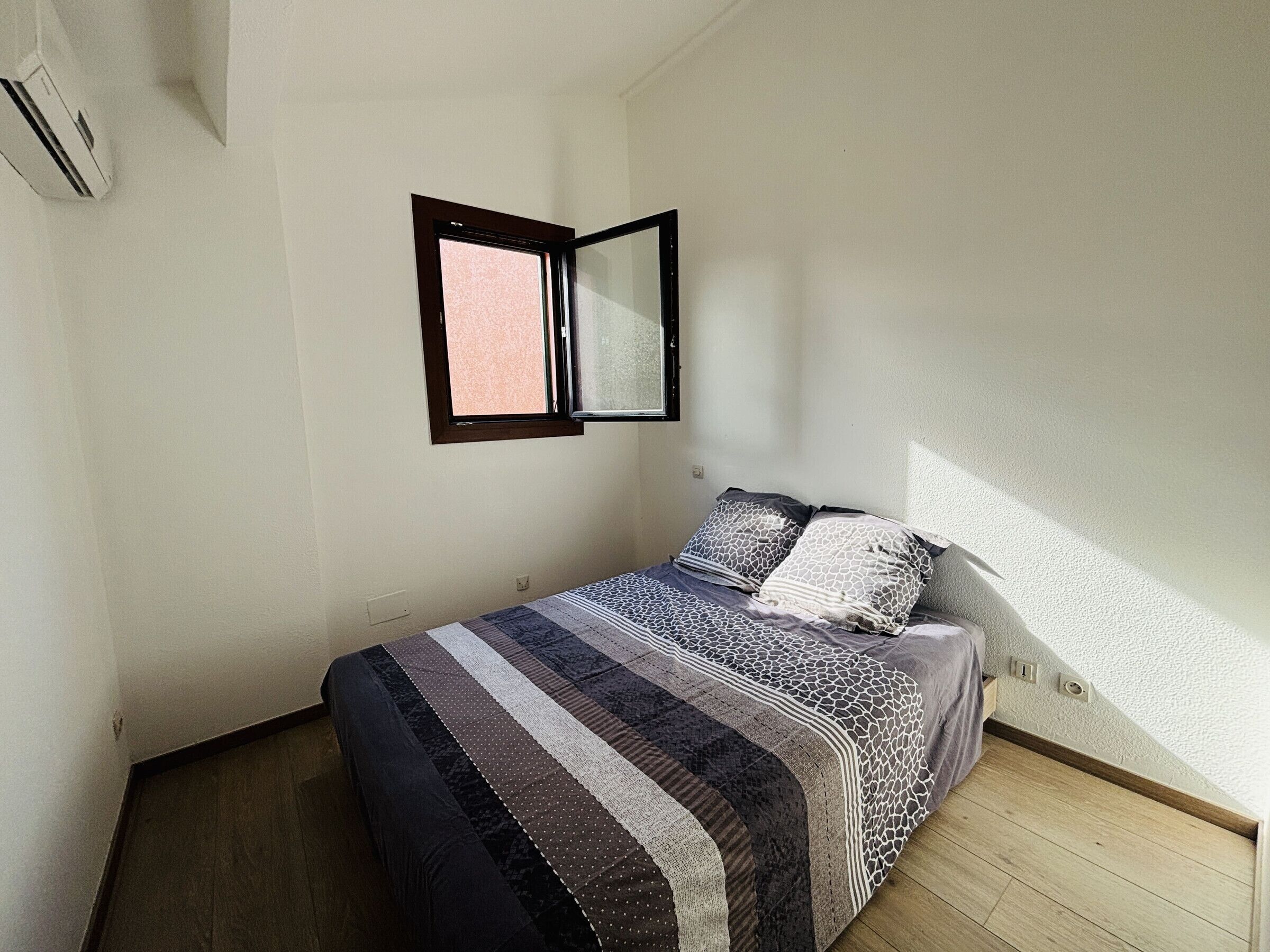 Una habitación pequeña con cama individual, ventana, suelo de madera y una colcha sencilla.