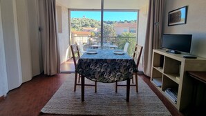 Ein Essbereich mit Tisch für zwei Personen, einem Fernseher auf einem Schrank und Blick auf eine Hügellandschaft durch eine Schiebetür.