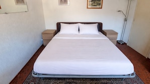Ein ordentlich gemachtes Bett in einem Hotelzimmer mit Kopfteil, Nachttisch, Stehlampe und zwei gerahmten Bildern an der Wand.