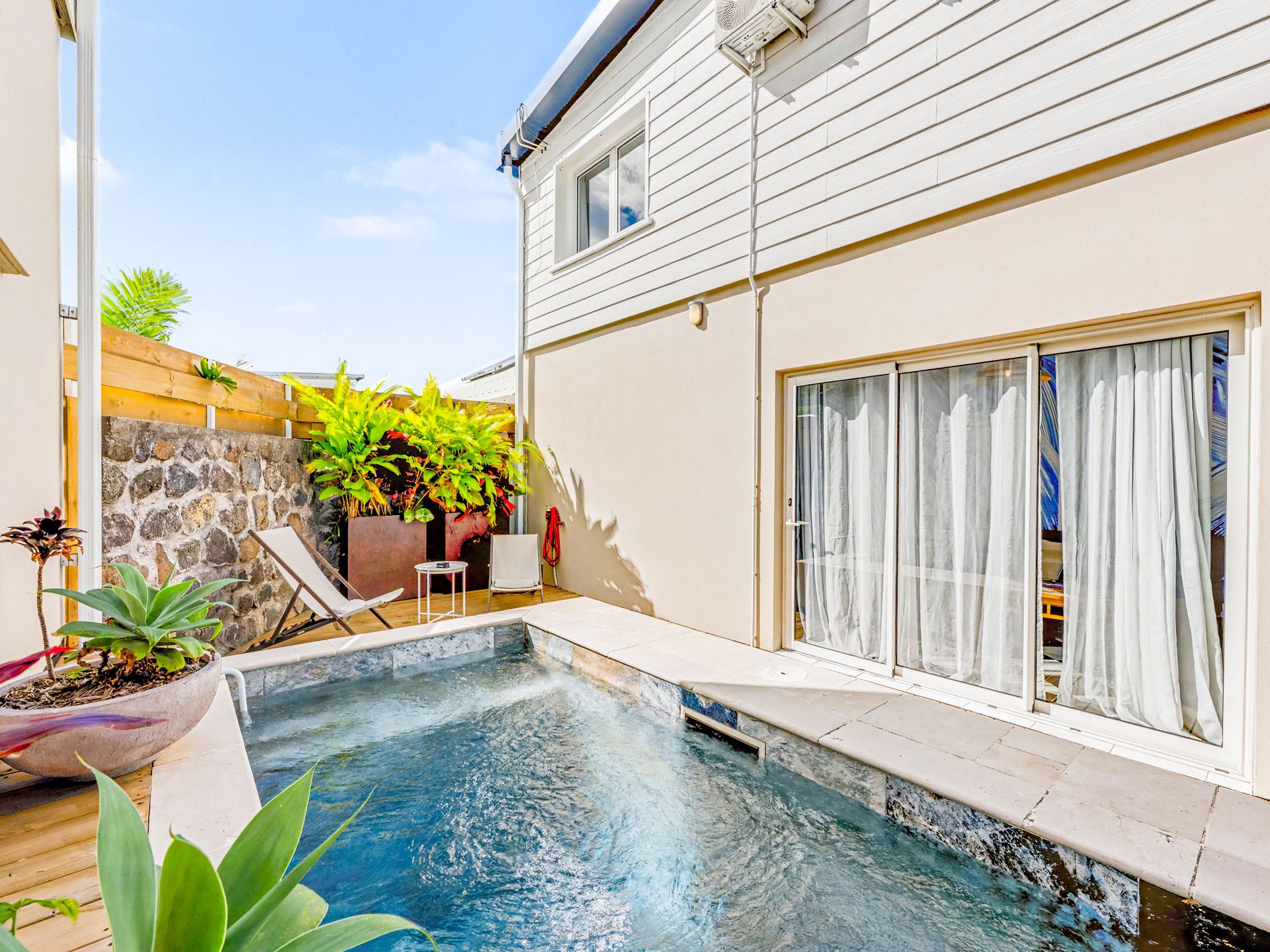 Una piscina con pared de piedra y plantas, terraza de madera y puerta corrediza de vidrio.