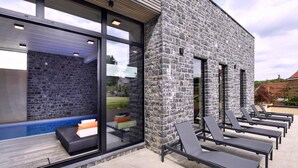 Un edificio moderno con paredes de piedra y amplias ventanas, sillones de exterior y una zona de piscina.