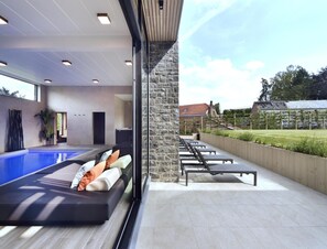 Een modern binnenzwembad met glazen wand, een loungegedeelte met een bank en kussens, en een stenen pilaar.
