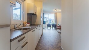 A narrow kitchen with a window, a sink, and cabinets. There is a chair and a table in the adjacent dining area. - Duinhoeve chalet Schelp (Burgh-Haamstede)