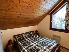 Ein Schlafzimmer mit schrägen Wänden, einem Bett mit kariertem Überwurf, einem Fenster mit Vorhängen und einem kleinen Nachttisch mit Lampe.