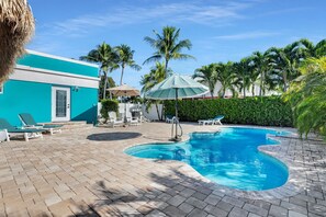 Ein Swimmingpool mit Liegestühlen und Sonnenschirmen, umgeben von Palmen und einem blauen Gebäude.