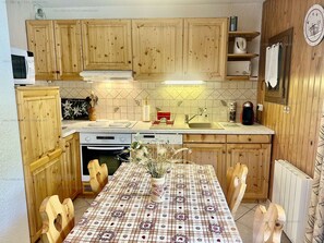 A kitchen with wooden cabinets, a microwave, a stove, a sink, and a dining table with a floral tablecloth. - Studio in village center - 4 guests (Valloire)