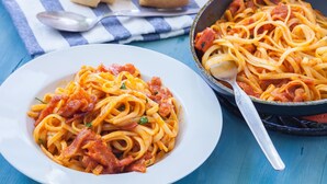 Un plato de espagueti con salsa de tomate y tocino, una sartén con más espagueti, y un mantel azul y blanco con pan y una cuchara sobre una mesa azul.