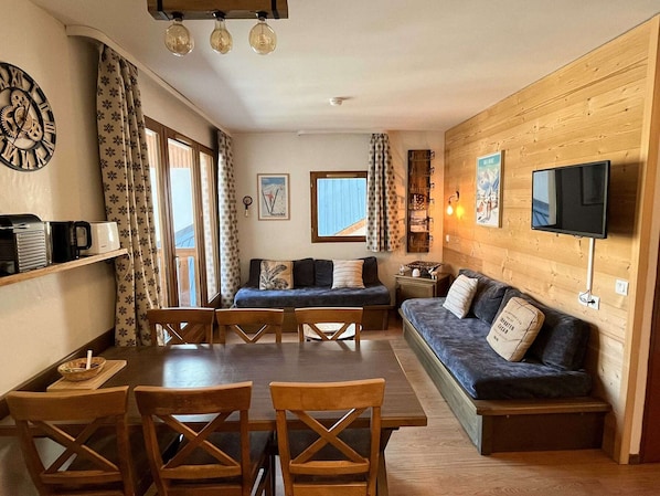 A cozy room with a wooden wall, a sofa, a TV, and a dining table with chairs.