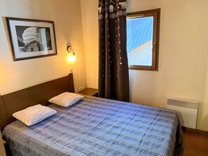 Une chambre d’hôtel comprenant un lit, une table de chevet, une lampe fixée au mur, un tableau encadré, une fenêtre avec des rideaux et un radiateur.