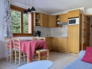 A compact kitchen with wooden cabinets, a dining table with chairs, a microwave, and a window with curtains. - Practical apartment near center - 6 guests (Valloire)