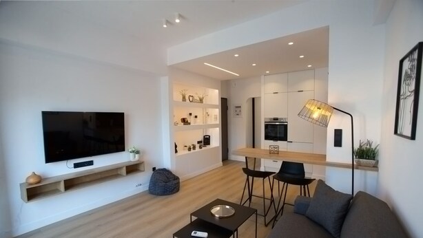 Un salon moderne avec un téléviseur à écran plat, un canapé, une table basse et un coin cuisine à l’arrière-plan.