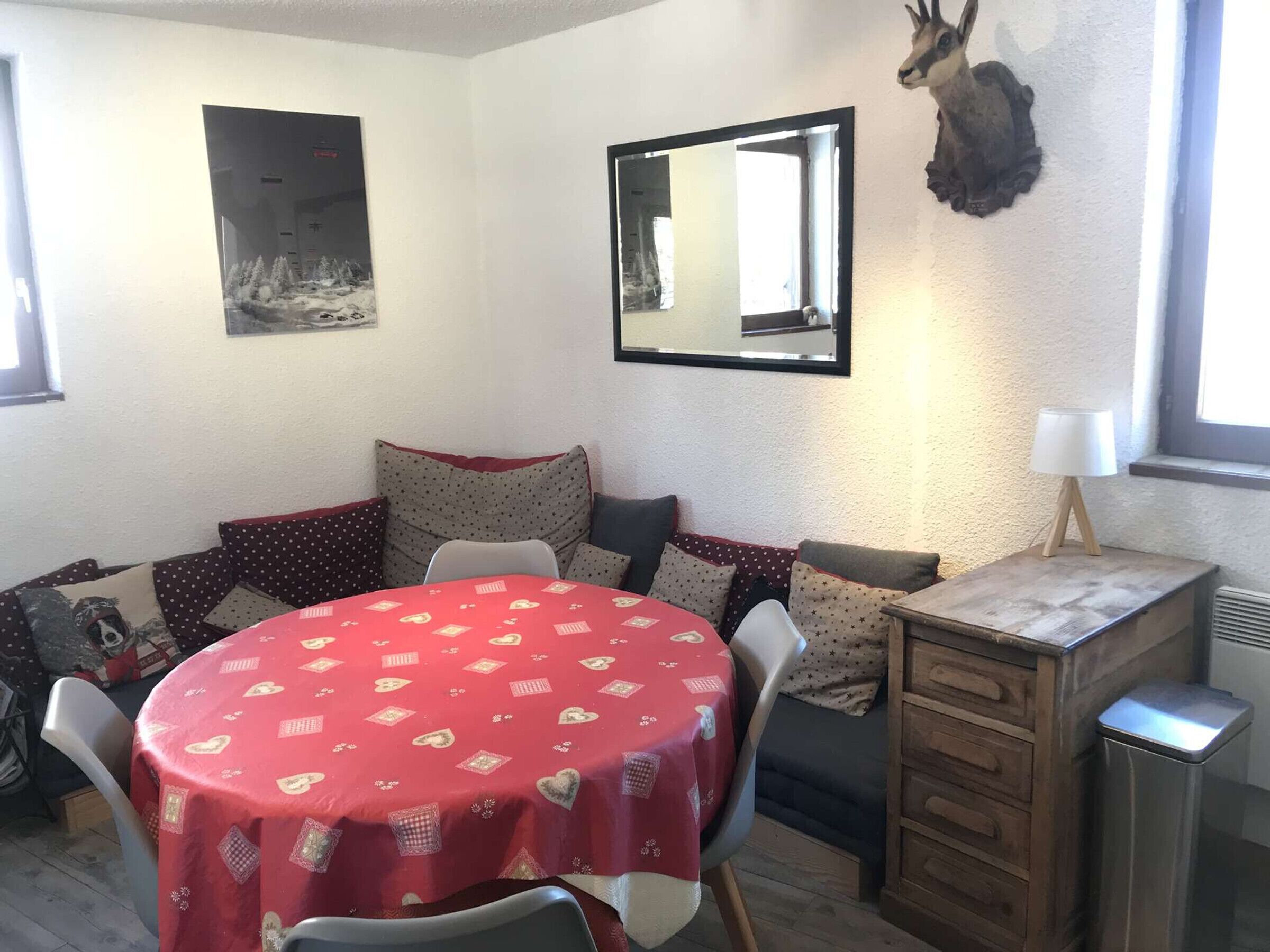 Ein gemütlicher Raum mit einem runden Tisch, der mit einer roten Tischdecke bedeckt ist, einer Couch mit gemusterten Kissen, einem Spiegel und einem aufgehängten Tierkopf an der Wand.