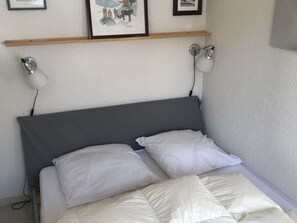Ein Bett mit grauem Kopfteil, weißen Kissen und einer weißen Daunendecke. An beiden Seiten des Bettes befinden sich Wandlampen.