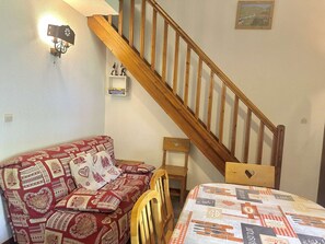 A cozy room with a wooden staircase, a patterned sofa, a small table, and a wall-mounted shelf with books. - Duplex apartment in Valloire - 6 guests (Valloire)