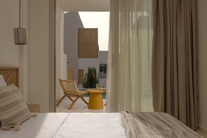 A modern hotel room with a bed, a small yellow table, a wicker chair, and a view of the outdoor area.