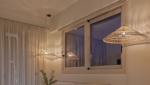 Un salon moderne avec un canapé, deux lampes suspendues, une fenêtre avec des rideaux et une petite table d’appoint en bois sur laquelle repose un magazine.