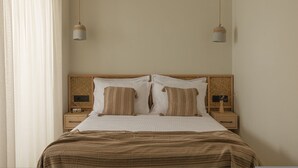 Ein ordentlich bezogenes Bett mit einer gestreiften Decke, zwei Nachttische mit Lampen und ein Fenster mit durchsichtigen Vorhängen.