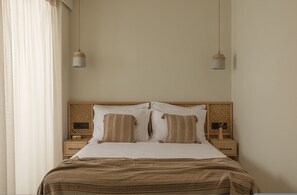 Ein ordentlich bezogenes Bett mit einer gestreiften Decke, zwei Nachttische mit Lampen und ein Fenster mit durchsichtigen Vorhängen.