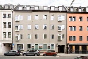 A row of modern apartment buildings with multiple floors, air conditioning units, and a parking area with cars.