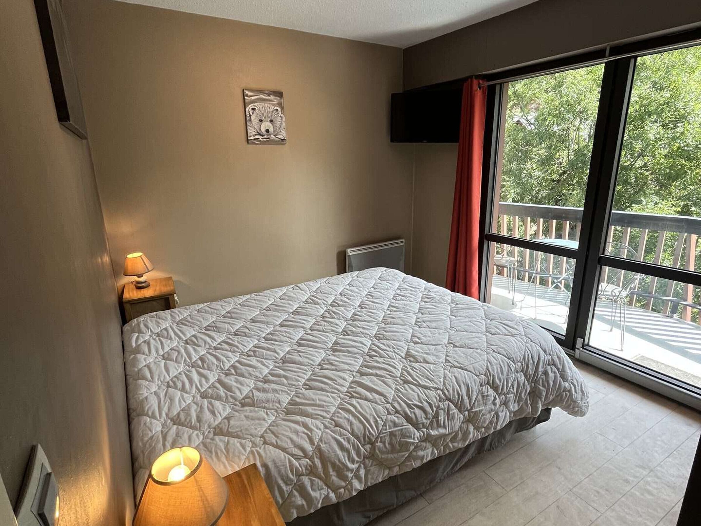 Ein Schlafzimmer mit Bett, Nachttischlampe, Fernseher und einem Fenster mit Blick ins Freie.