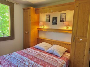 Een slaapkamer met een houten hoofdbord, twee bedlampen, een bed met beddengoed met een patroon, en een raam met uitzicht op bomen.