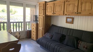 A room with a wooden cabinet, a sofa, a table, and a window with a view of greenery. - Cozy studio cabin - 4 guests (Valloire)