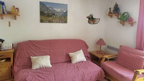 A cozy living room with a red patterned sofa, a wooden side table, a lamp, a wall-mounted duck decoration, and a framed mountain landscape picture. - Studio in the center - pets welcome - 4 guests (Valloire)