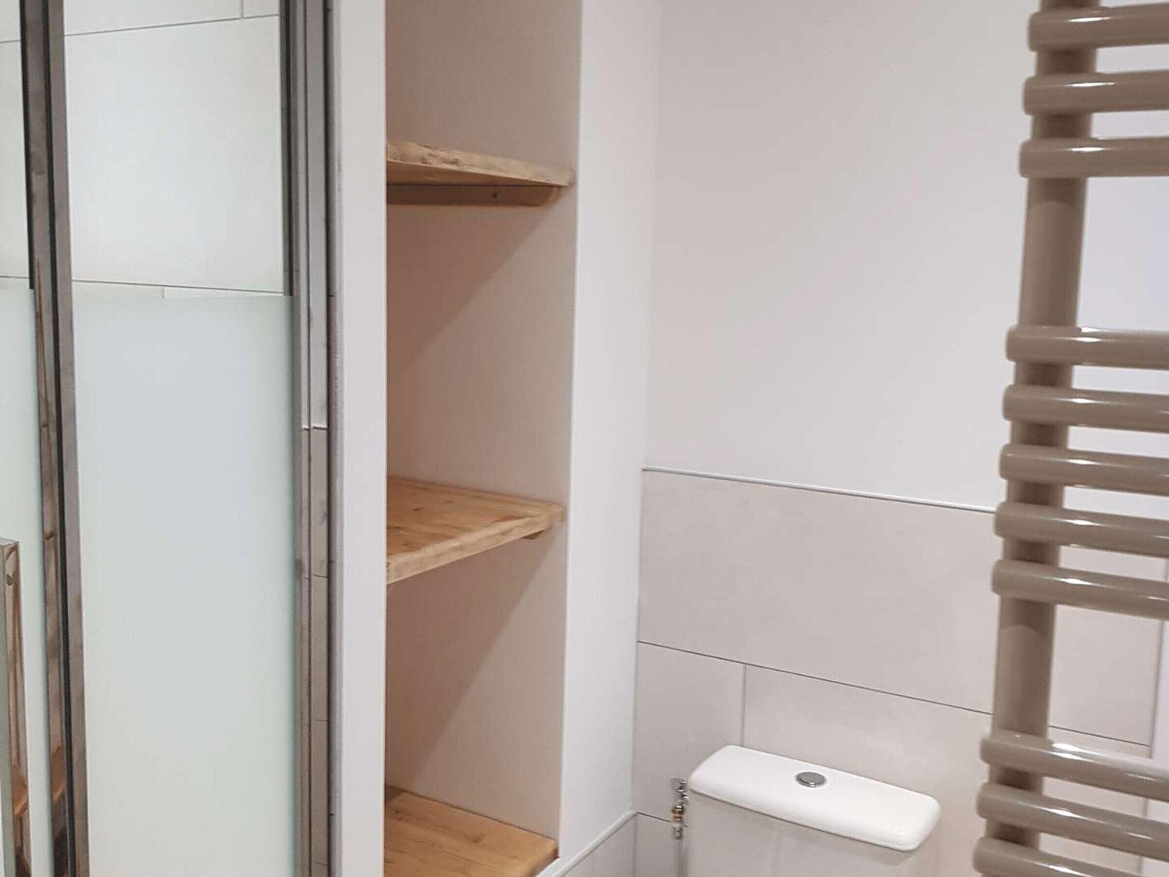 A bathroom with white tiled walls, a wooden shelf, and a toilet with a white flush handle.