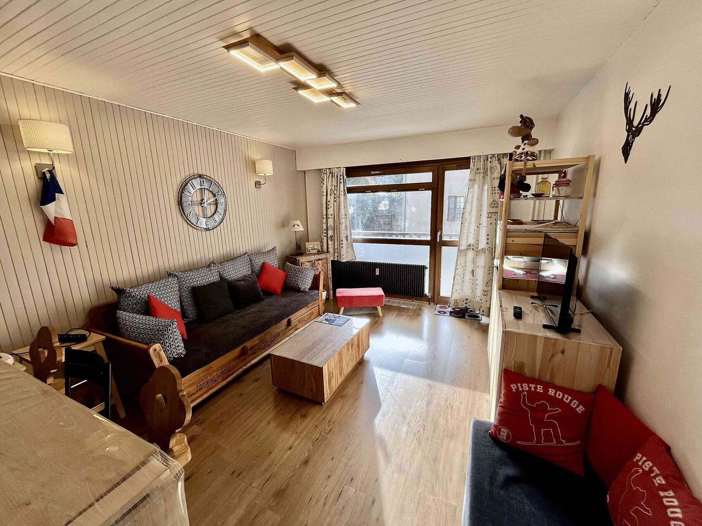 A living room with a sofa, coffee table, and a bookshelf. There is a window with curtains, a radiator, and a wall clock.