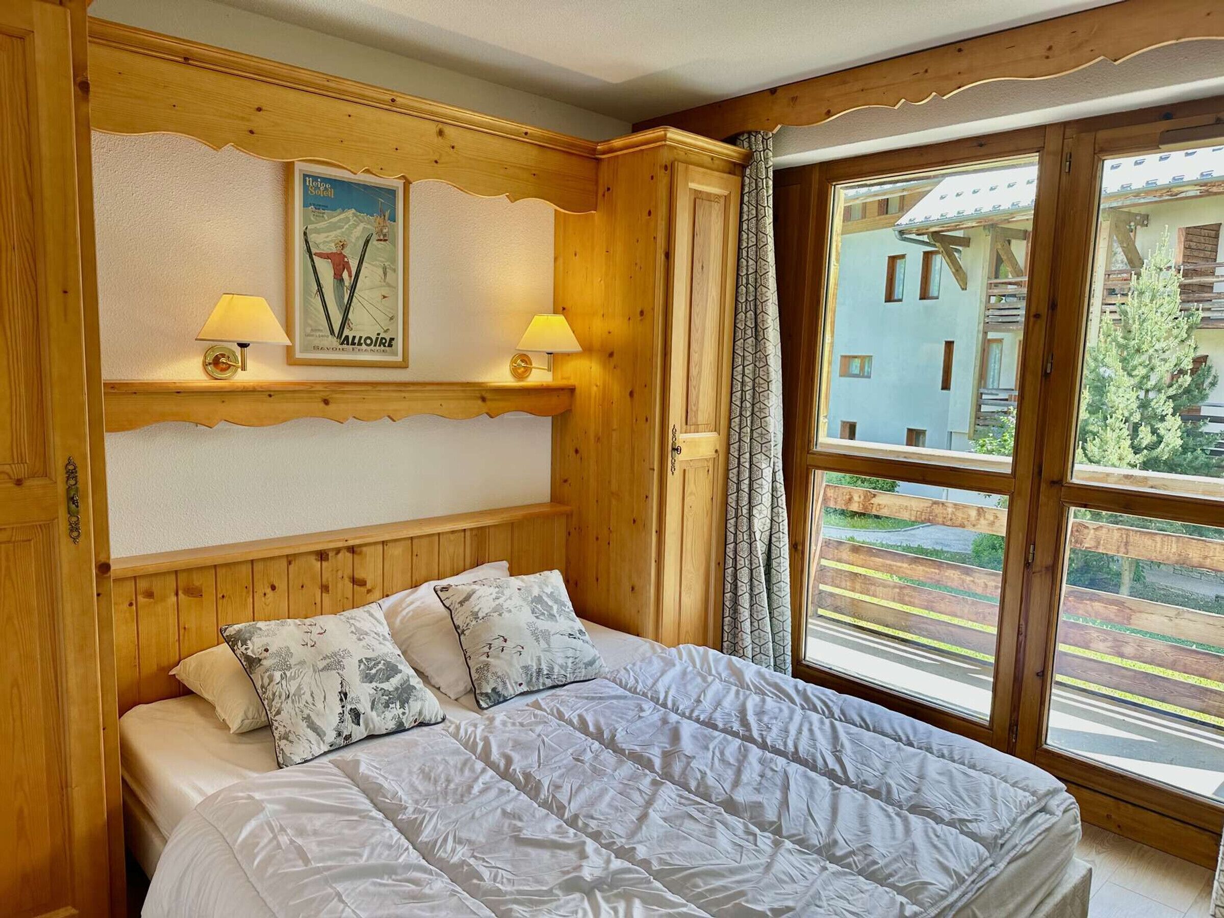 Una habitación de madera con una cama, dos lámparas, un cuadro enmarco y una ventana que ofrece vistas a edificios y árboles.