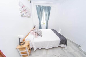 A bedroom with a bed, a nightstand, a lamp, a window with curtains, and a wall-mounted painting. - My Sevillian Retreat (Seville)