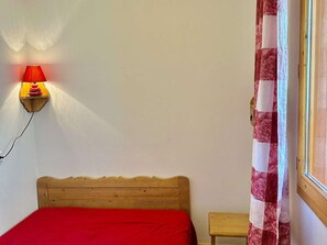 A room with a wooden bed, a red bedspread, a wall-mounted lamp, and a window with a floral curtain. - Cozy apartment with elevator - 4 guests (Valloire)