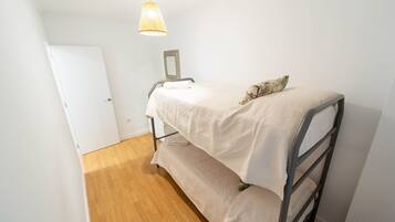 Una habitación pequeña con cama individual, piso de madera, puerta blanca y una lámpara colgante.