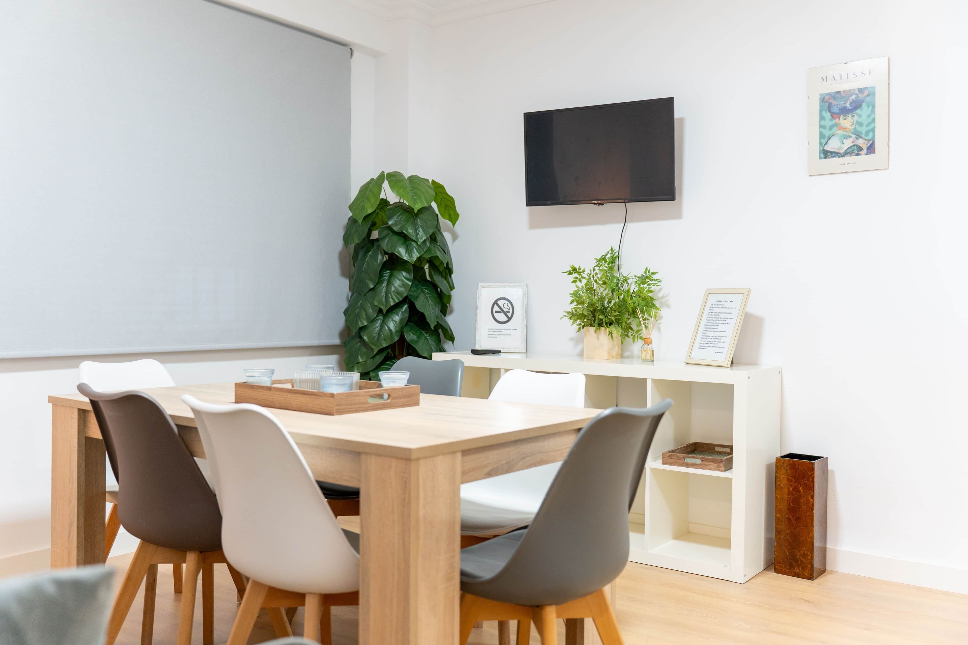 A modern meeting room with a wooden table, white chairs, a large green plant, a mounted TV, and a white shelving unit with a framed certificate.
