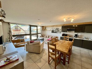 A spacious living area with a sofa, a dining table with chairs, a kitchen area, and a large window offering a view of a building with a sloped roof. - Comfortable apartment with parking - 6 guests (Valloire)