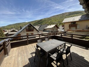 A wooden deck with a table and chairs, overlooking a village with traditional houses and a hill in the background. - Comfortable apartment with parking - 6 guests (Valloire)