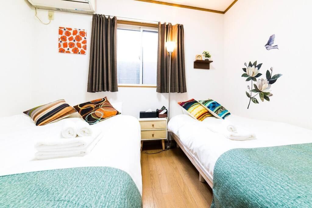 A hotel room with two beds, a window with curtains, a wall-mounted air conditioner, a small shelf with a plant, and a wall decoration of flowers and birds.
