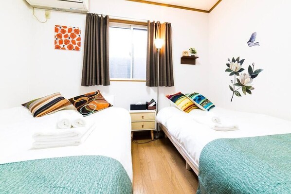 Une chambre d’hôtel avec deux lits, une fenêtre avec des rideaux, un climatiseur fixé au mur, une petite étagère avec une plante et une décoration murale représentant des fleurs et des oiseaux.