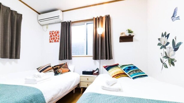 A hotel room with two beds, a window with curtains, an air conditioning unit, a small shelf with a plant, and wall art. - Honmachi 2 Chome (Tokyo)