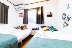 Ein Hotelzimmer mit zwei Betten, einem Fenster mit Vorhängen, einer Klimaanlage, einer kleinen Ablagefläche mit einer Pflanze und Wandbildern.