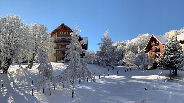 A snow-covered landscape with two wooden buildings, one with a balcony and another with a sloped roof. - Elegant apartment with parking - 6 guests (Valloire)