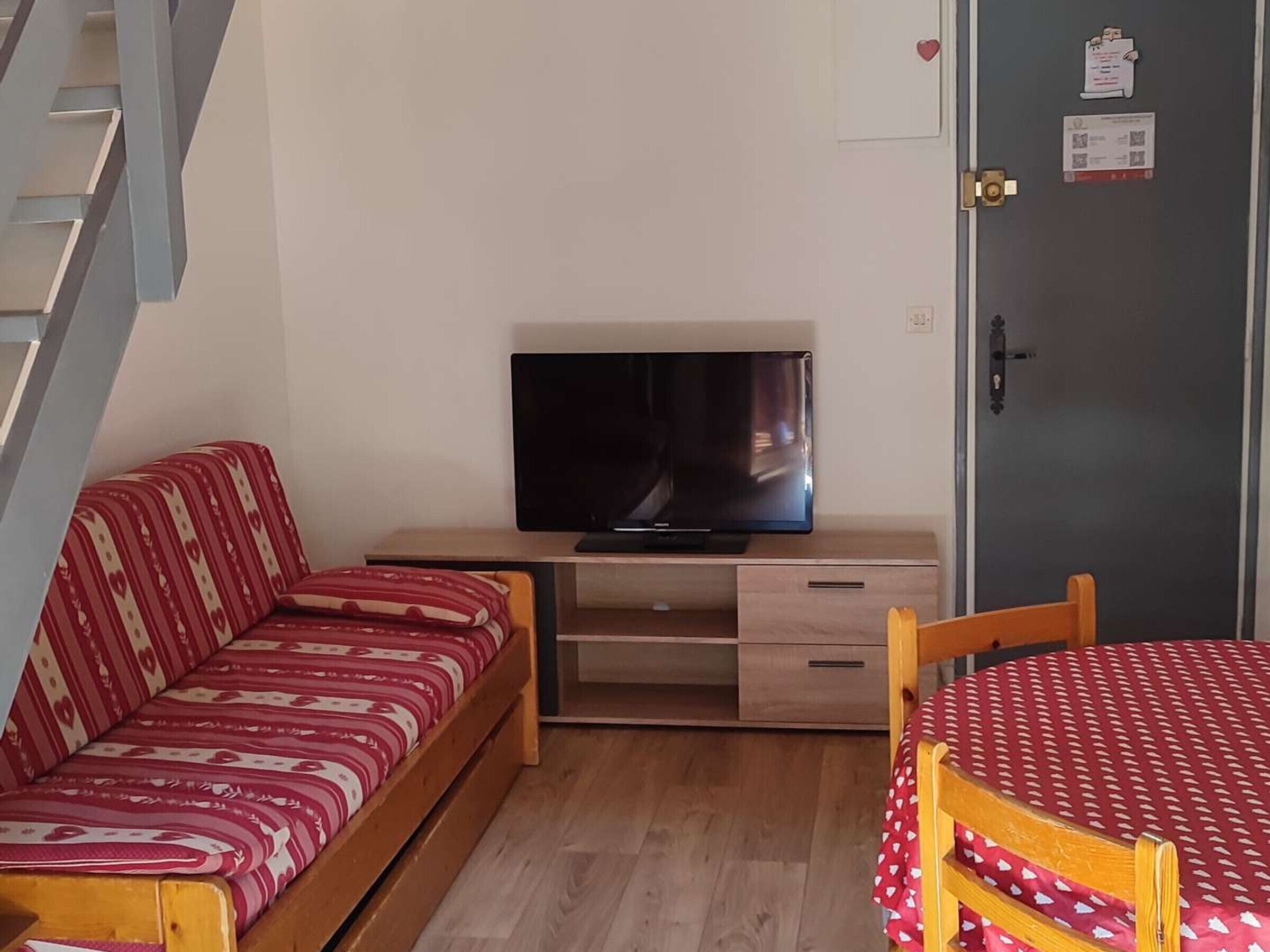 Een kamer met een bank, een televisie op een houten standaard, een tafel met een rood-wit geruit tafelkleed, en een deur met slot.