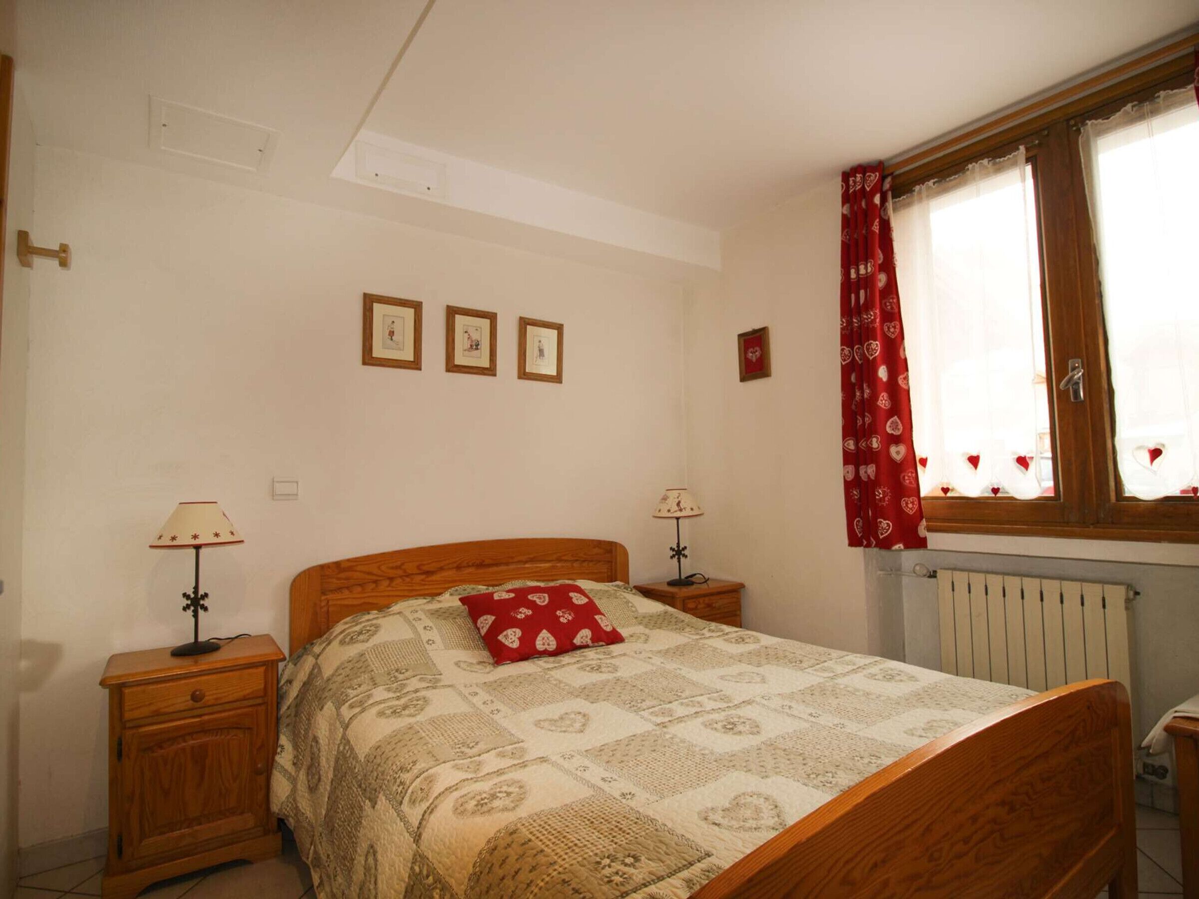 Une chambre à coucher avec un lit en bois, deux tables de chevet avec des lampes, un radiateur et une fenêtre avec des rideaux rouges.